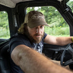 A man with a beard driving a vehicle, wearing a brown trucker hat with 'The Element' written on it.