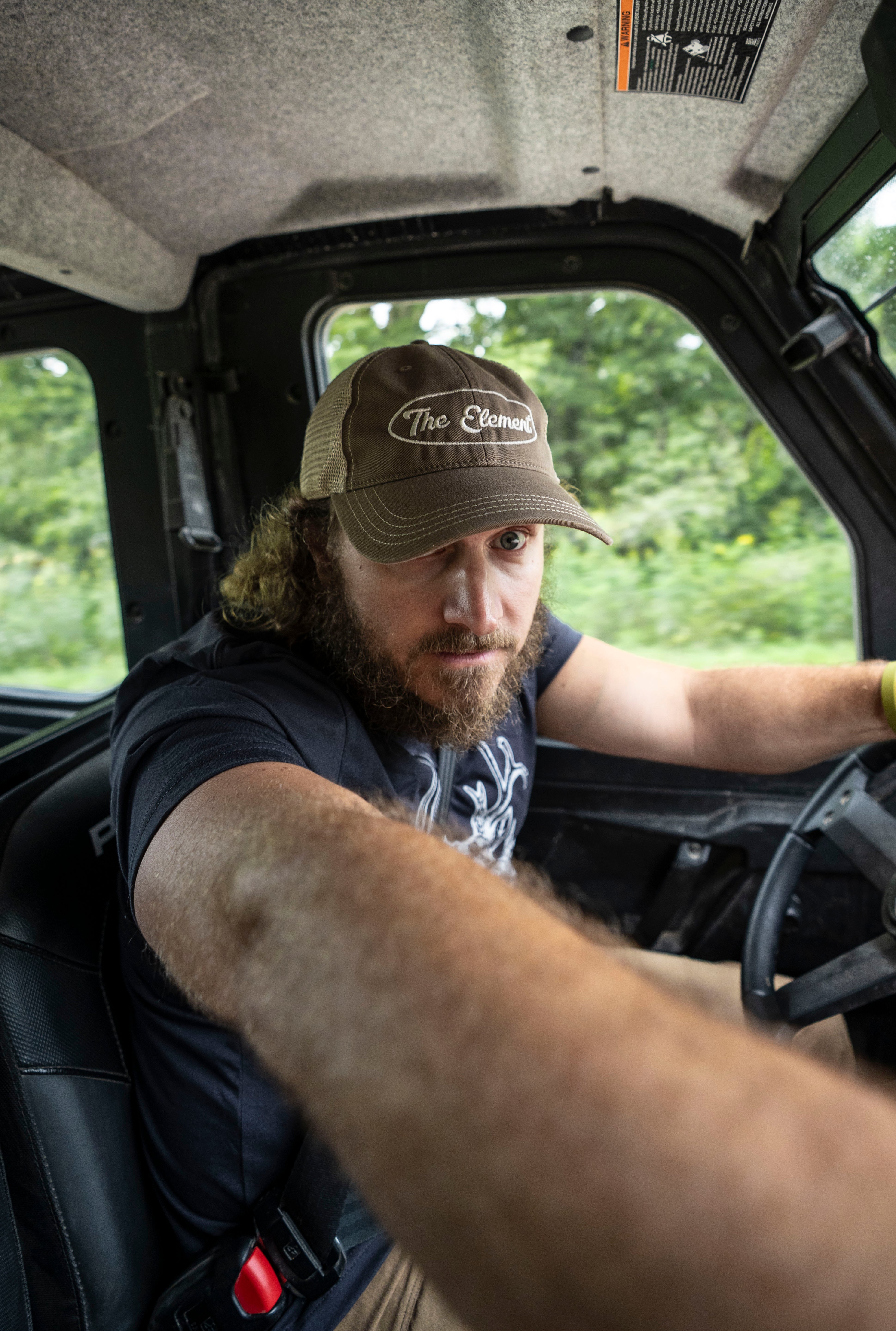 A man with a beard driving a vehicle, wearing a brown trucker hat with 'The Element' written on it.